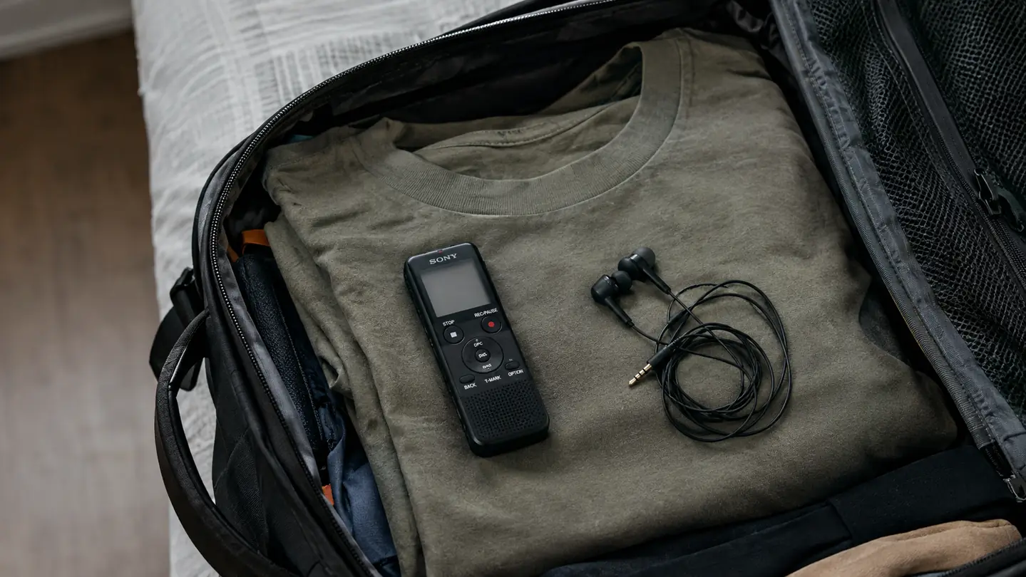 A small portable audio recorder and a pair of earbuds lying on a folded t-shirt inside an open carry-on backpack