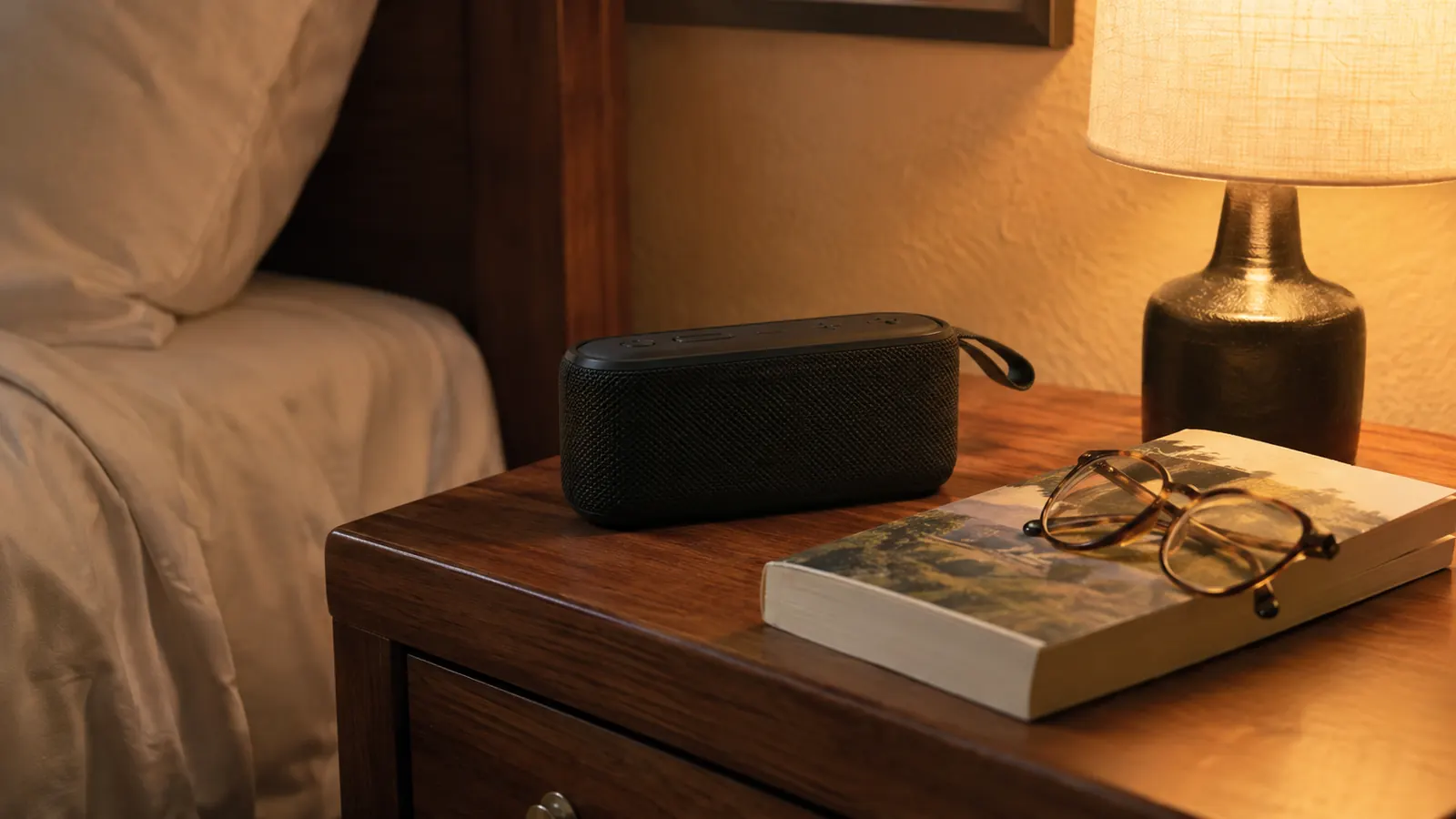 A portable speaker standing upright on a motel nightstand beside a paperback book and reading glasses