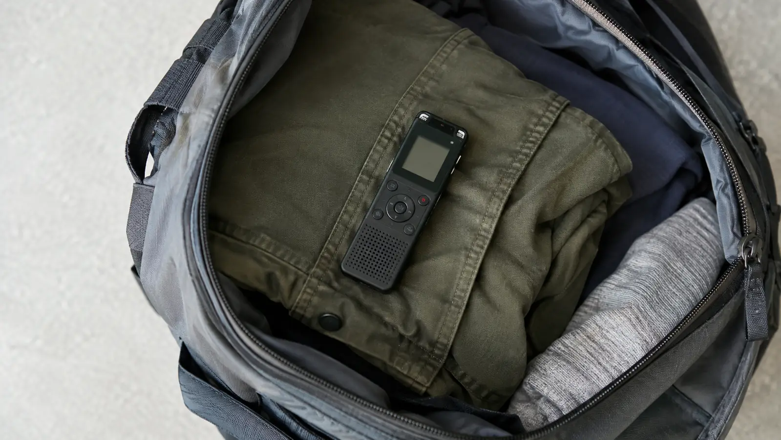 A small handheld recorder lying on a folded jacket inside an open carry-on backpack